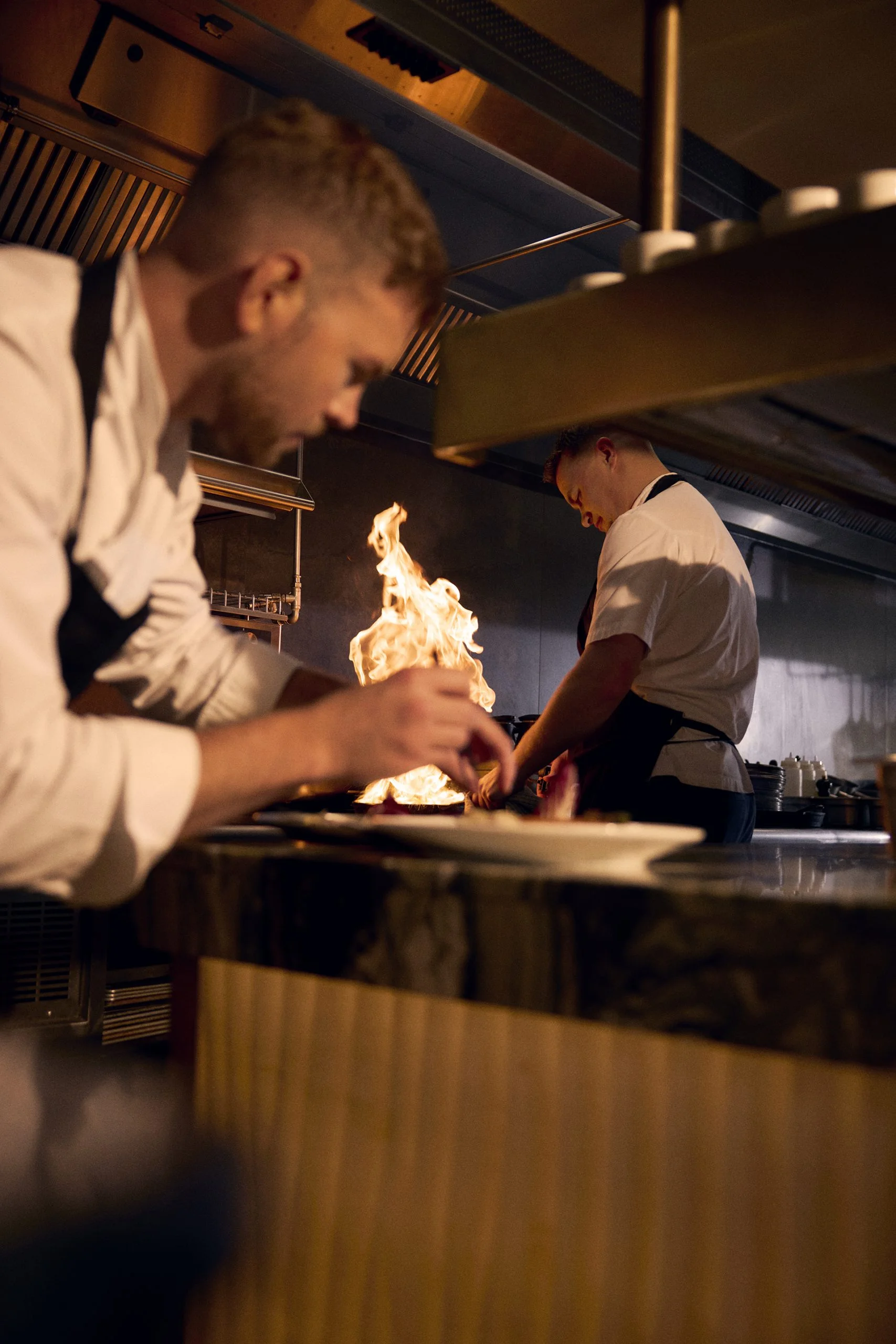 man plating food and fire behind him in a kitchen